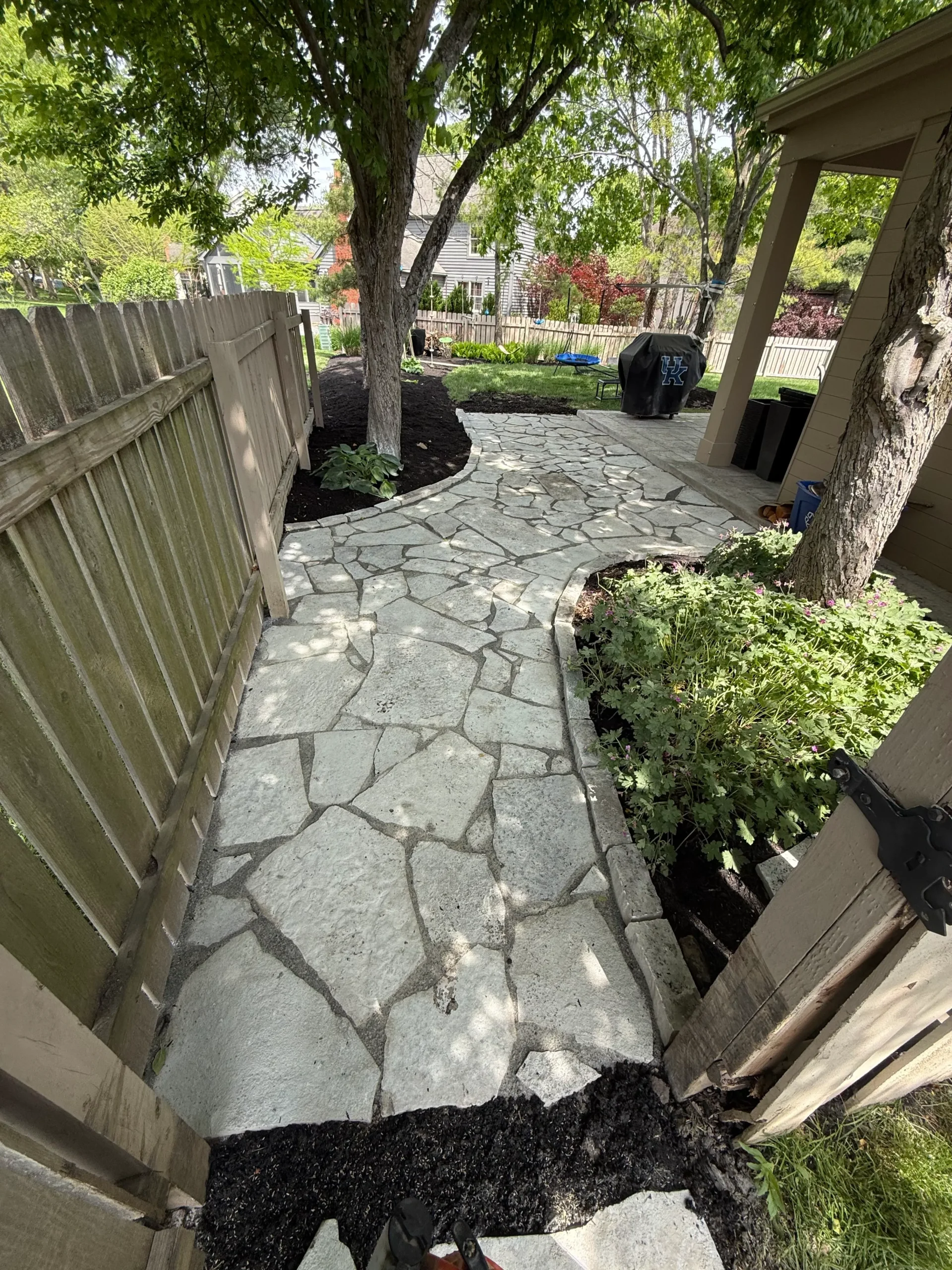 Stone flagstone path curves through a shaded backyard with a wooden fence and trees, and a covered grill in the distance.