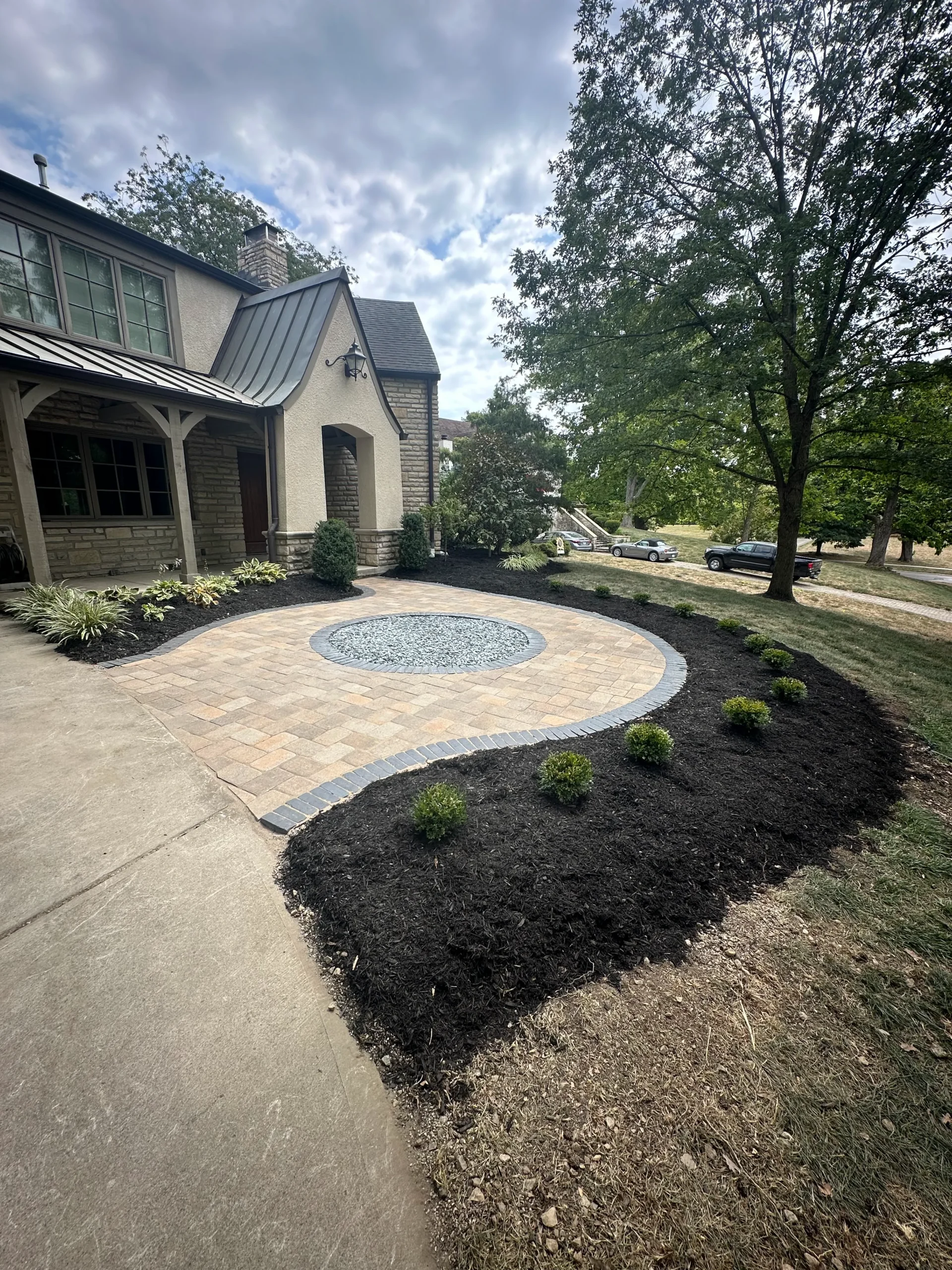 Front of a stone house with a circular paved courtyard and a pebble-filled centerpiece, surrounded by mulch and small shrubs.